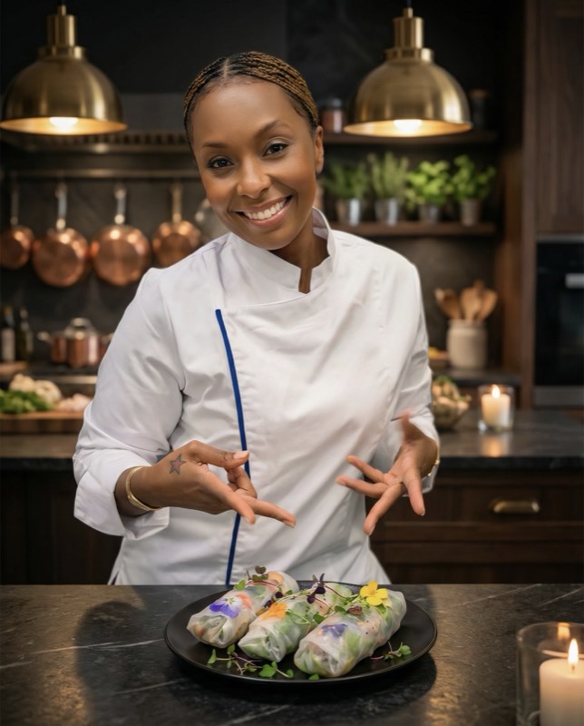 Chef Ashè in chef&rsquo;s whites in a dark kitchen, presenting a plate of fresh spring rolls with edible flowers.
