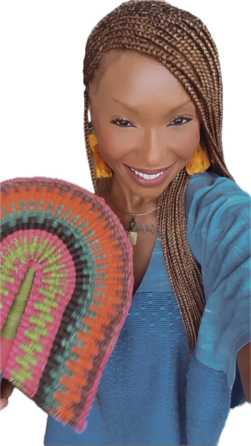 Chef Ashé smiling warmly, with long honey-brown braids, yellow tassel earrings and a teal top, holding a colourful woven fan.