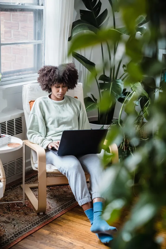 A woman in a sage sweatshirt connecting online from a laptop, curled in a chair among leafy houseplants.