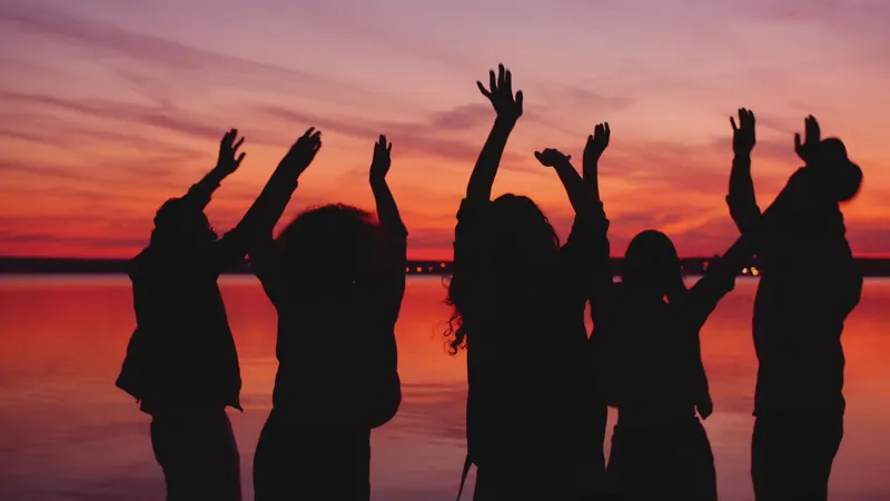 Silhouettes of friends dancing with arms raised against a glowing orange and violet sunset over water.