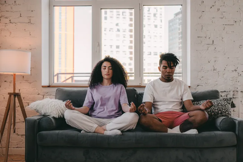 A couple seated cross-legged on a sofa, eyes closed, meditating side by side in a sunlit loft.