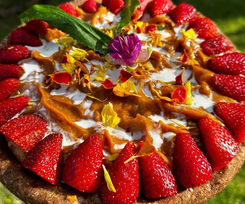 A raw Sunfired tart crowned with strawberries, cashew cream and edible marigold petals, glowing in the sun.