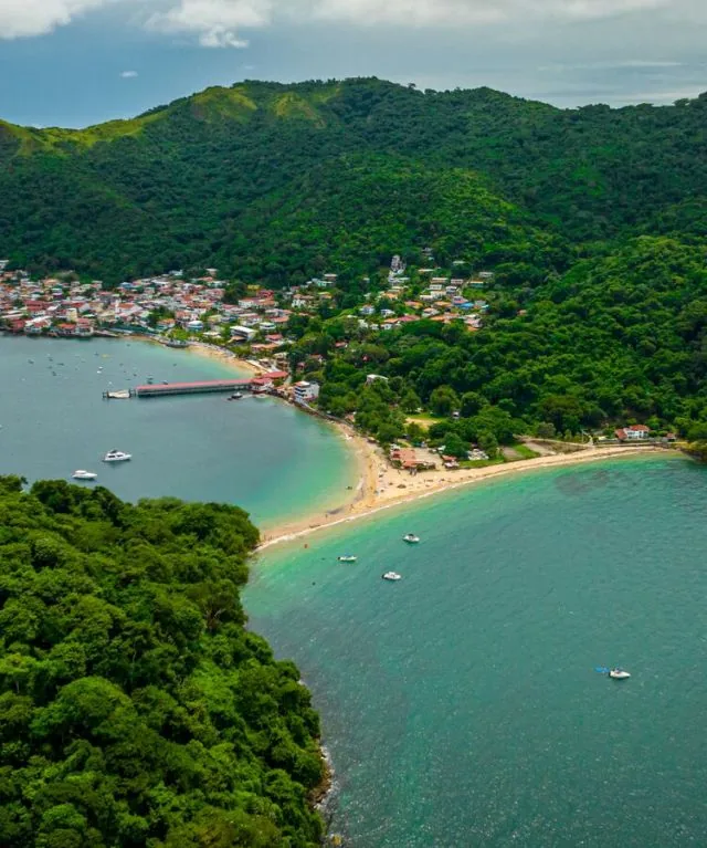 Aerial view of Taboga: a curved golden beach and turquoise bay below a hillside village wrapped in green forest.