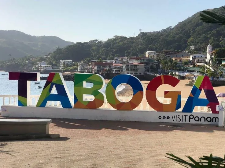 Colourful TABOGA letters on the seafront promenade, with the beach, village and green hills behind.